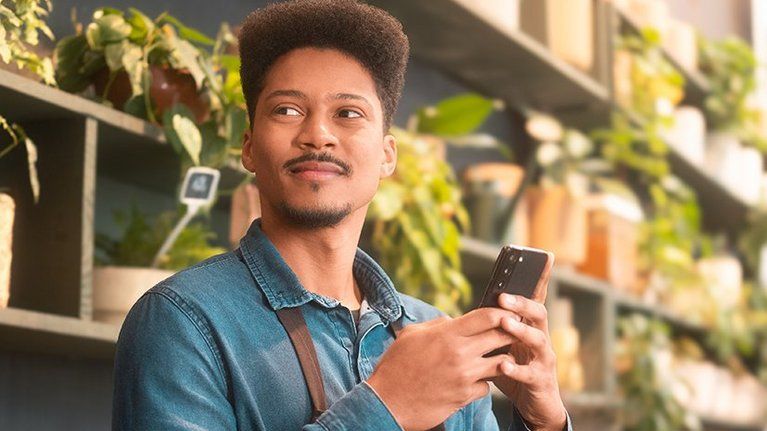 A smiling man holds a smartphone in front of shelves filled with green plants.